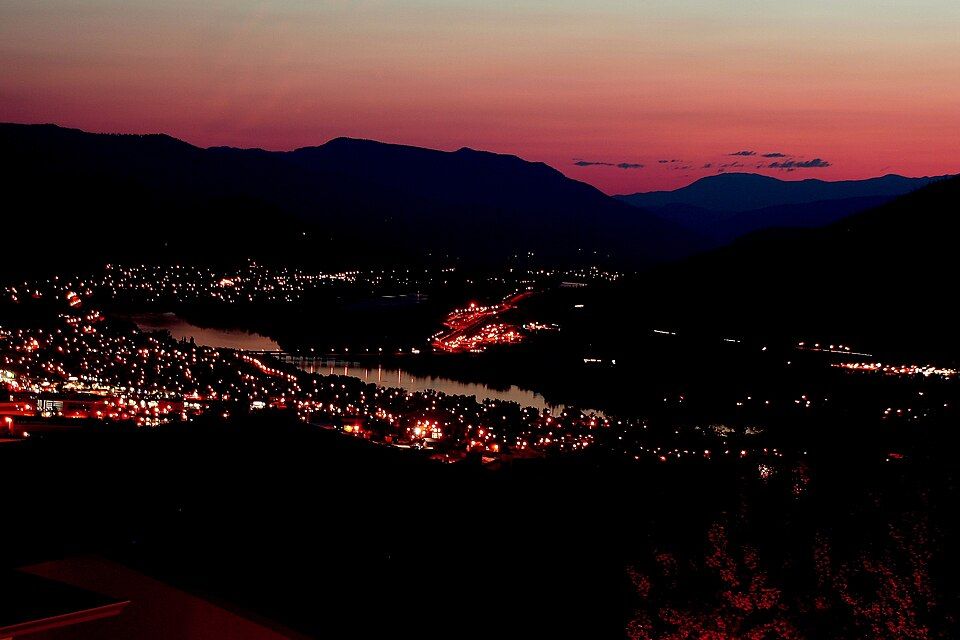 A town still lit up at night beside a river, under a pink pre-dawn sky over the mountains.