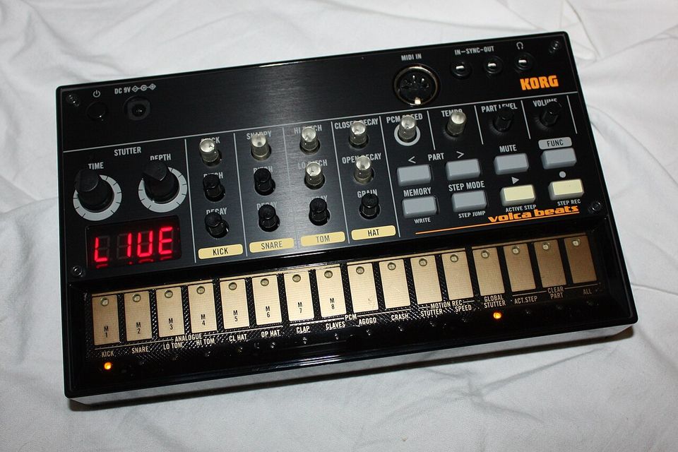 Korg Volca Beats lying on a white cloth with LIVE on its display, showing the knobs, part buttons and sixteen step keys.
